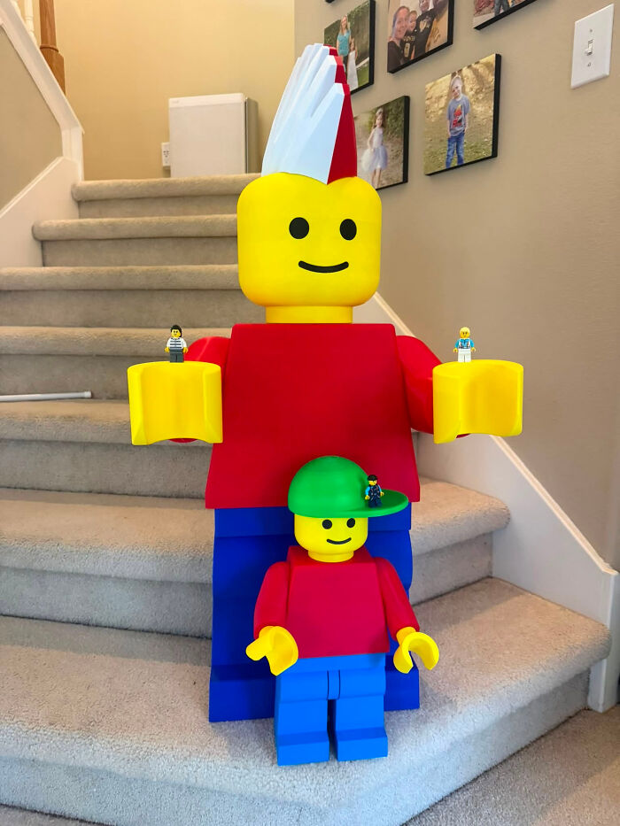 Two oversized 3D printed Lego figures, a parent and a child, standing on a staircase, with mini Lego figures in their hands and on their hat.