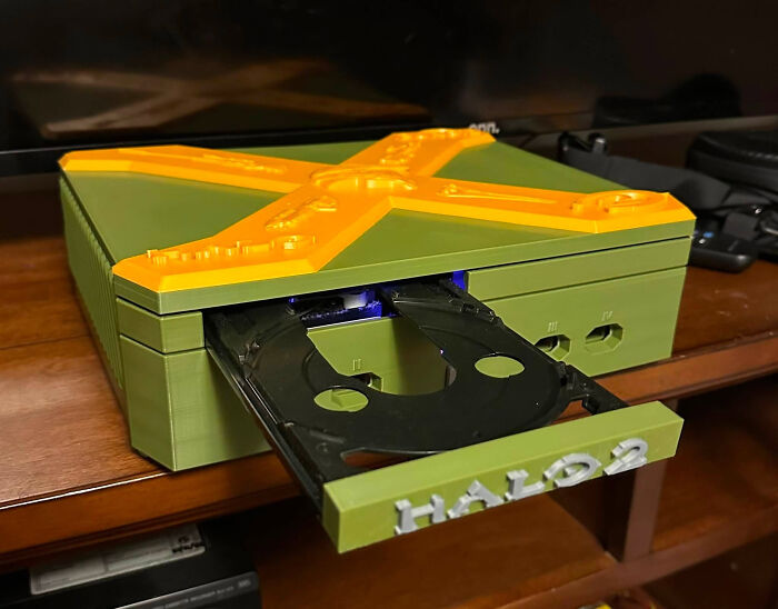 A custom 3D printer case for an Xbox, in green and orange, with the disc tray open revealing Halo 2 text.