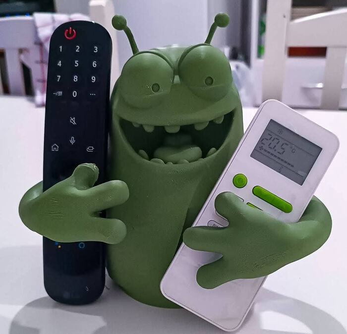 A quirky green monster figure, created with a 3D printer, holding two remote controls.