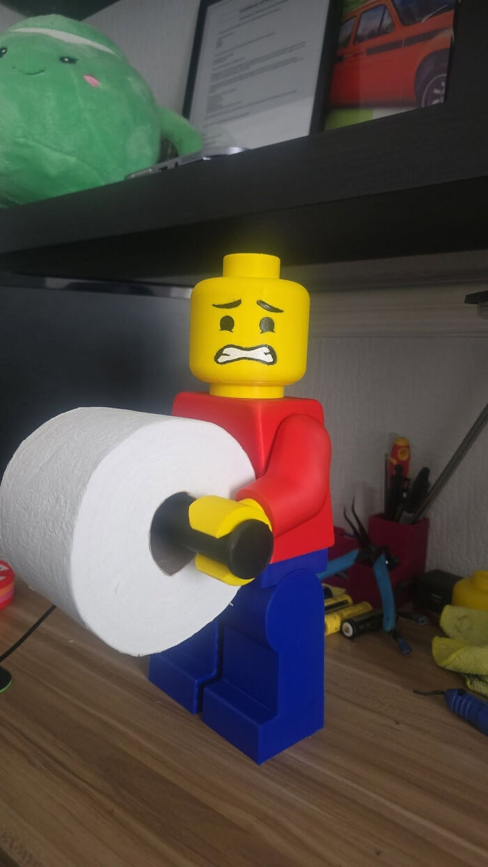 A large 3D printer Lego figure with a distressed expression, holding a roll of toilet paper.