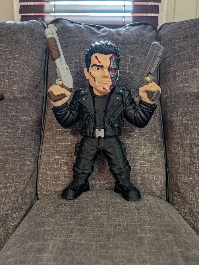 A detailed 3D printer creation of a Terminator action figure holding two guns, seated on a gray couch.