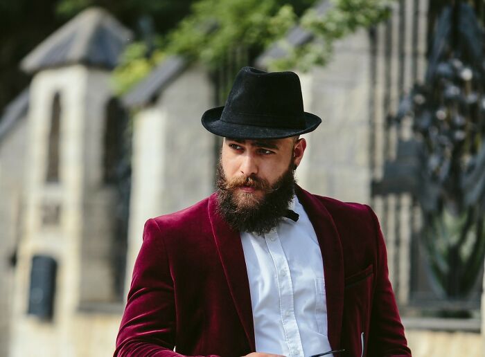 A man with a beard, black hat, and red velvet blazer looks to the side outdoors. Products, Trends, and Items.