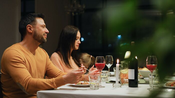 A couple at a candlelit dinner, with the man looking content and the woman looking away, perhaps about a boyfriend's gift.