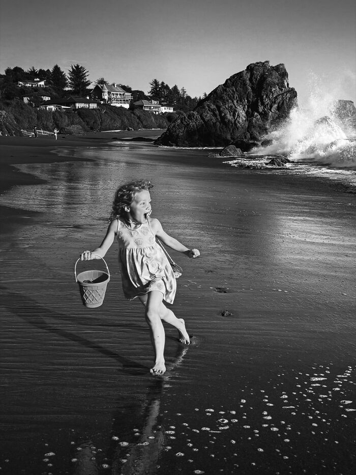 A young girl joyfully runs on a beach with a bucket, a black and white image from the iPhone Photography Awards.