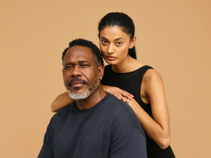A serious looking age-gap relationship couple, an older man and younger woman, pose against a neutral background.