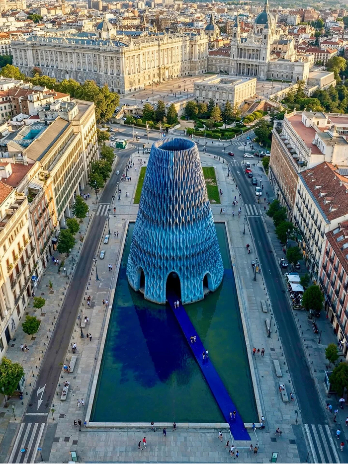 An aerial view of an amazing modern architecture design featuring a blue, textured conical building surrounded by a reflective pool in a city.