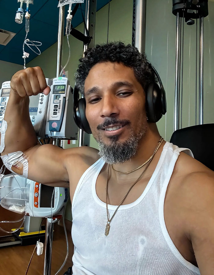 A man needing a transplant, flexing his bicep while connected to medical equipment, smiling and wearing headphones.