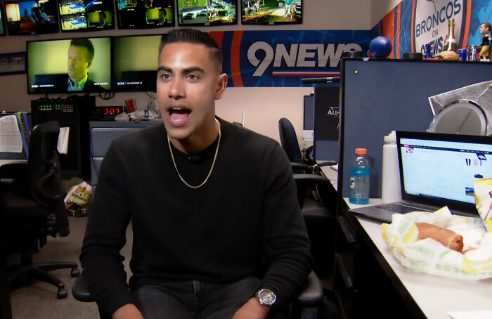 A man in a black shirt and gold chain sits in an office, speaking with an open mouth, possibly discussing an alleged cheater.