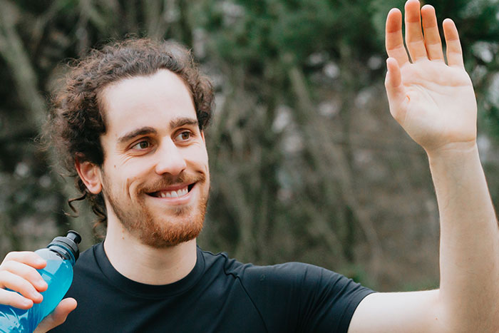 A smiling man with curly hair, holding a blue sports drink and waving, reflecting on unfortunate names.