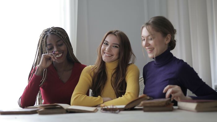 Three young women smiling and looking at the camera, with open books in front of them, discussing a boyfriend's gift.