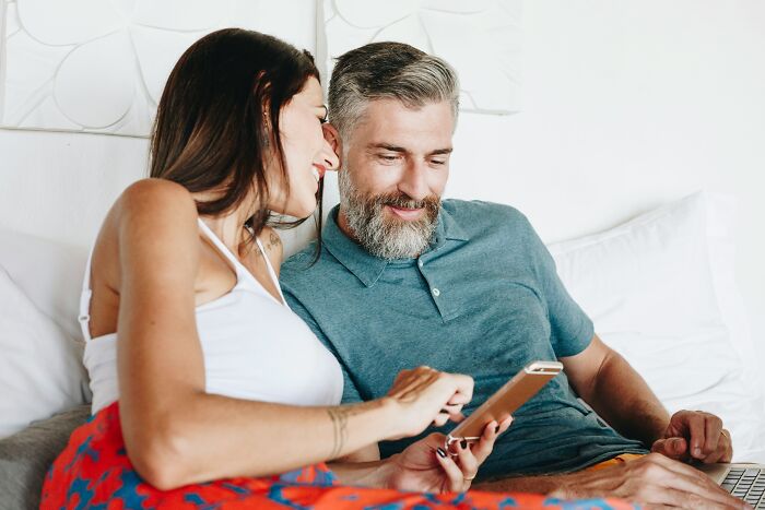 A mature man with a beard and a younger woman looking at a phone, sharing moments in their age-gap relationship.