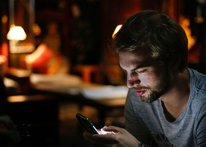 A man intensely looks at his phone in a dimly lit room. B*m-maxxing drives men toward their worst selves.