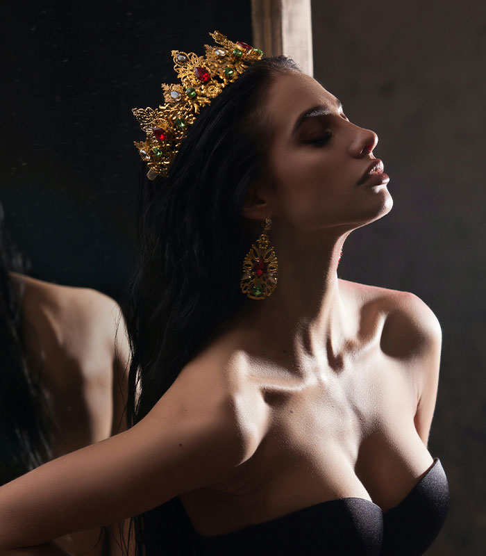 A woman wearing a gold crown and elaborate earrings, symbolizing the empowering intimate position of Queening. A woman wearing a gold crown and elaborate earrings, symbolizing the empowering intimate position of Queening.