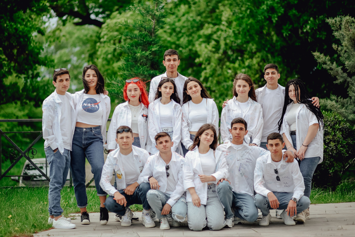 A group of young students in white shirts and jeans smiling outdoors, evoking a mysterious atmosphere.