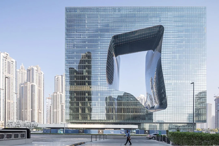 A striking modern architecture design of a glass-clad skyscraper with a large, fluid-shaped void in its center, reflecting the sky and surrounding buildings.