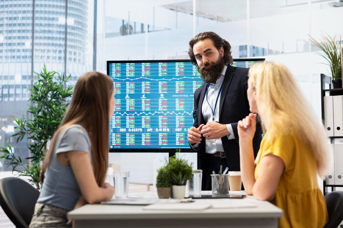 A manager with a beard explains stock data to two women in an office, illustrating dumb reasons people get fired.