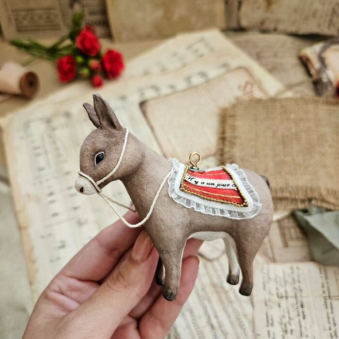 A handmade cotton wool donkey creation with a red and white saddle, held against old paper and red flowers.