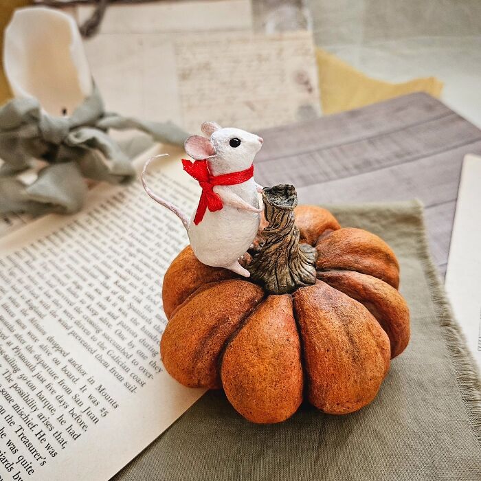 A handmade cotton wool mouse creation wearing a red scarf, standing on an orange pumpkin against vintage papers.
