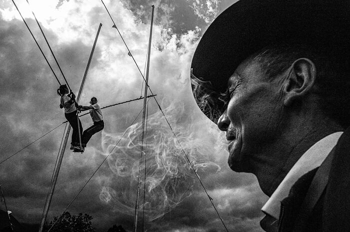 A man in a hat watches two acrobats on a swing against a cloudy sky. A dynamic black and white photograph.