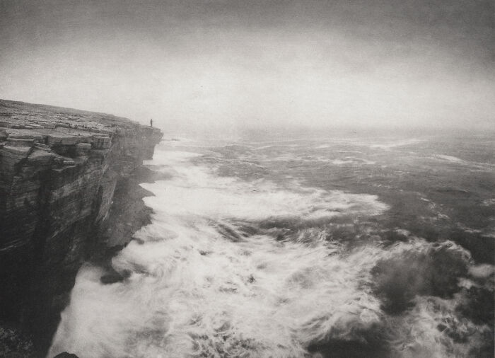 A lone figure stands on a cliff overlooking a rough, stormy sea. An award-winning black and white photograph.
