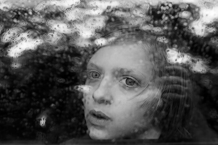 A young woman's face, partially obscured by water droplets on a window, in a captivating black and white photograph.