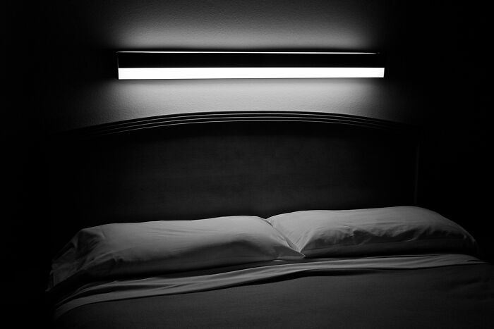 A dark room with a bed and illuminated light fixture overhead. A stunning black and white photograph.
