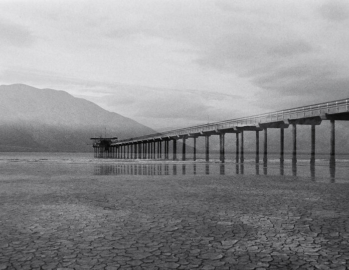 Long pier extends over cracked earth and water in a captivating black and white photograph.