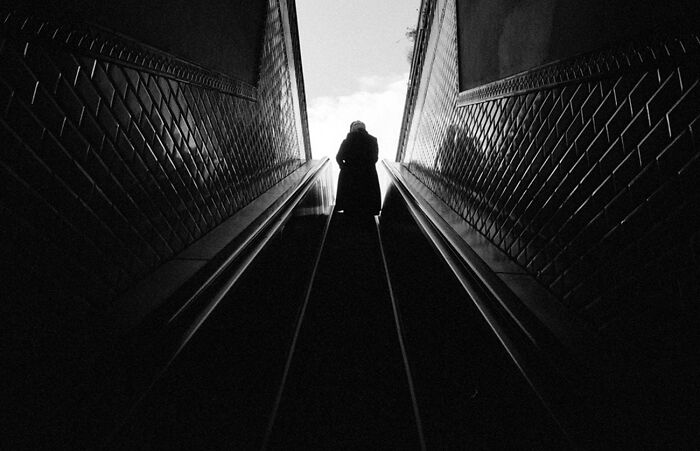 Dramatic black and white photograph of a lone figure ascending an escalator towards a bright sky.