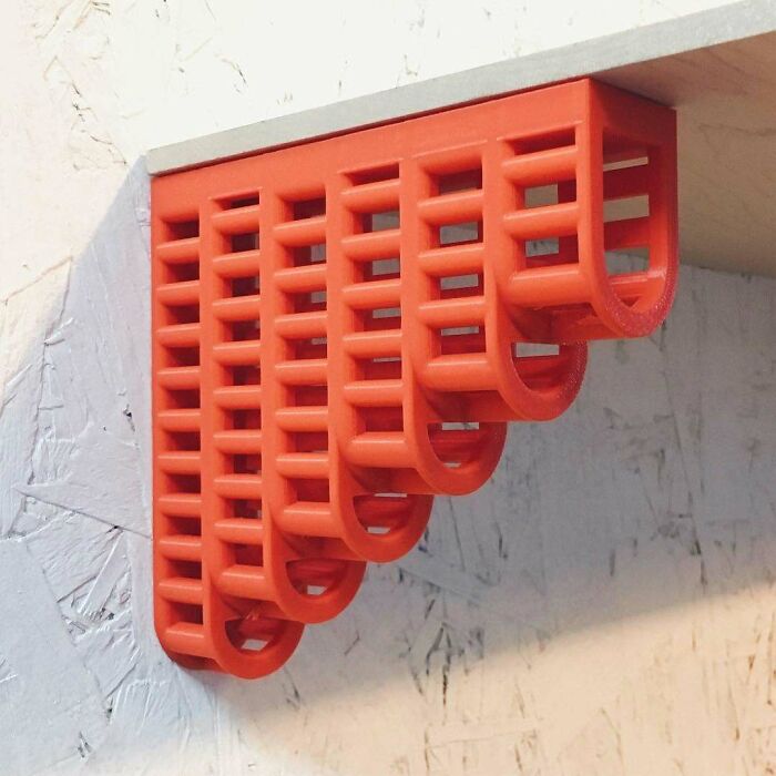 An orange 3D printing shelf bracket with a unique lattice design, securely mounted on a white wall.