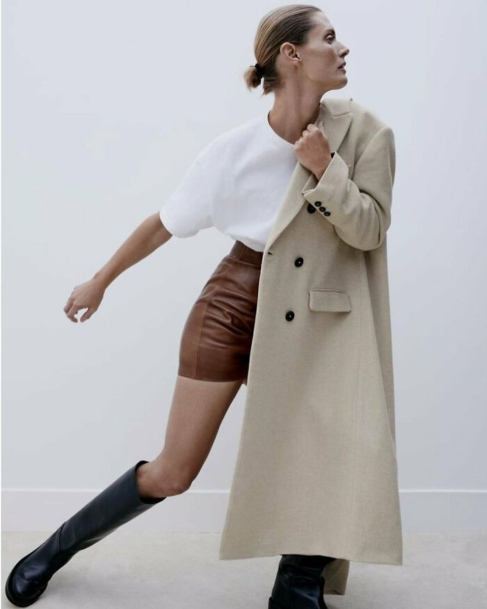 A model in bizarre Zara content wearing a white t-shirt, brown leather shorts, a long beige coat, and black boots.