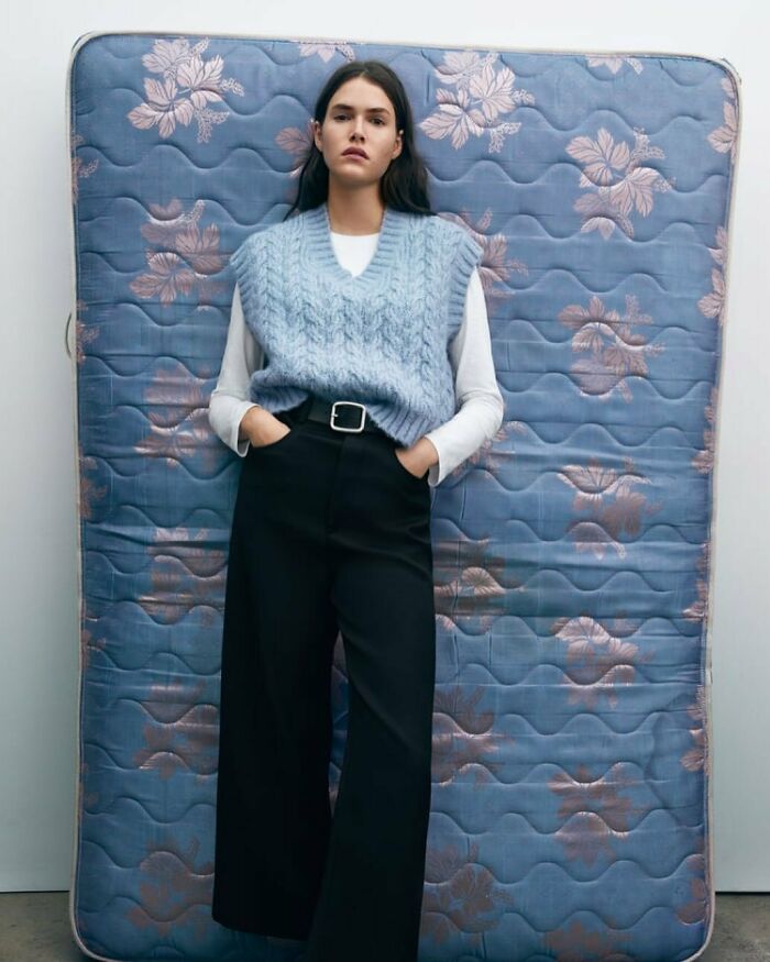 A woman in a blue sweater vest posing against a mattress, from the bizarre content found on Zara website.
