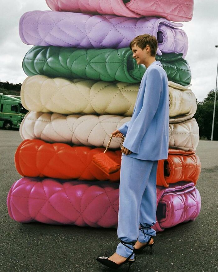 A woman in a light blue suit stands next to a tall stack of colorful, quilted bags, illustrating bizarre Zara content.