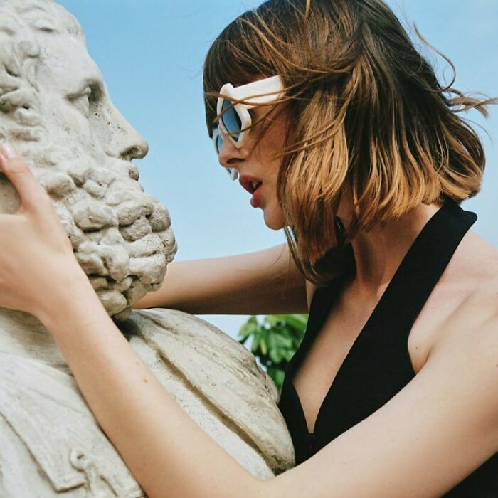 A woman wearing stylish sunglasses closely examining a stone bust, highlighting bizarre content on the Zara website.
