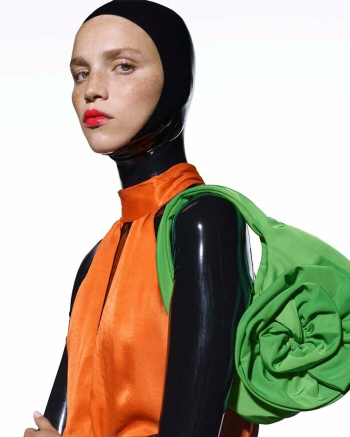 Model in bizarre content from Zara, wearing a black hood, orange top, and carrying a green bag.