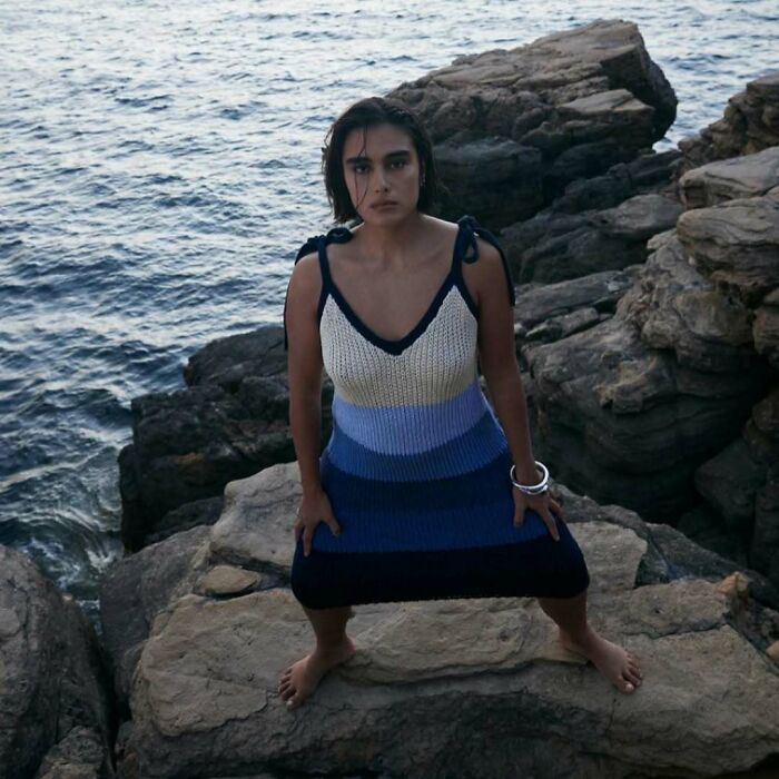A woman in a striped knit dress poses on rocks by the ocean, part of Zara's bizarre content.