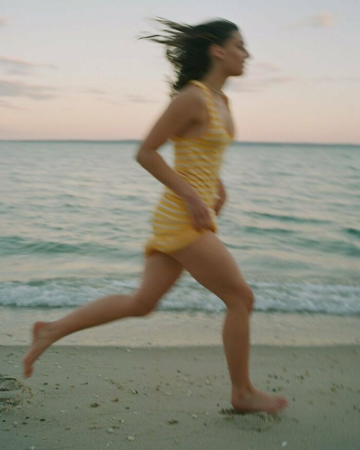 A woman in bizarre Zara content runs barefoot on a beach at sunset, wearing a striped yellow dress.