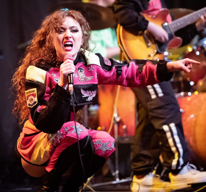 A female celebrity with curly red hair, holding a microphone and pointing, embodying the spirit of disillusioned celebrities.