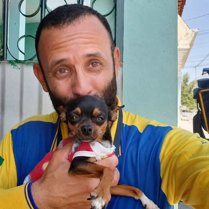 A mailman with a beard holds a tiny dog in a red and white outfit, showcasing his sweet bond with dogs.