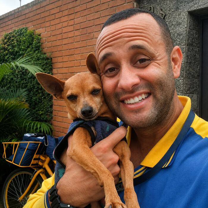 A cheerful mailman holds a small brown dog in a sweater, demonstrating his kind interactions with dogs during deliveries.