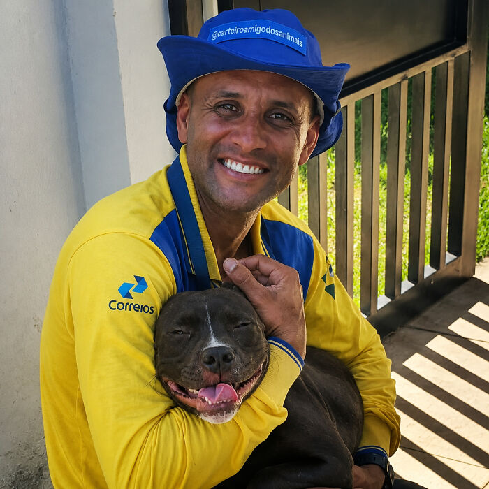 A happy mailman in a blue hat with text hugs a content black dog, highlighting his heartwarming befriending of dogs.