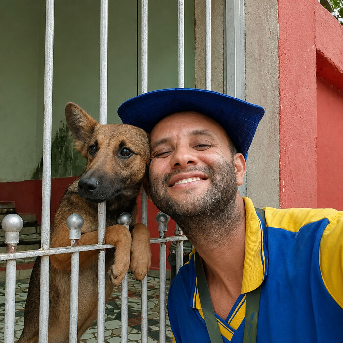 A kind mailman leans against a fence, smiling with a dog peeking through the bars, stopping to befriend dogs.