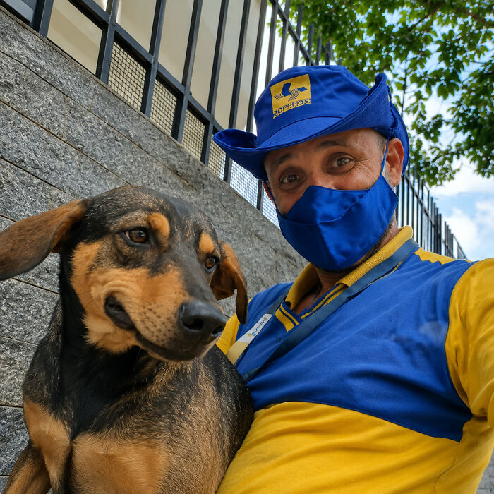 A kind mailman in uniform with a blue face mask poses with a happy brown dog, stopping to befriend dogs.