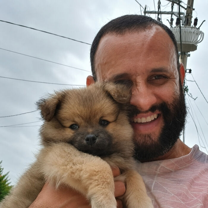 A mailman smiles widely, holding a small fluffy brown puppy, highlighting his heartwarming interactions with dogs.