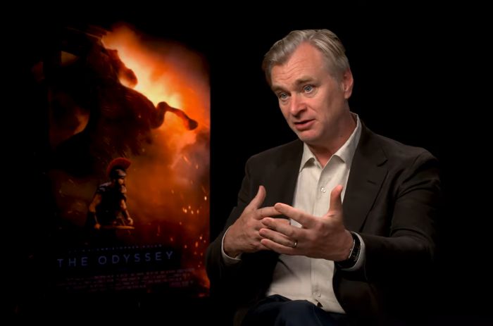 A man speaking in front of a movie poster for The Odyssey. This image is related to The Odyssey film adaption.