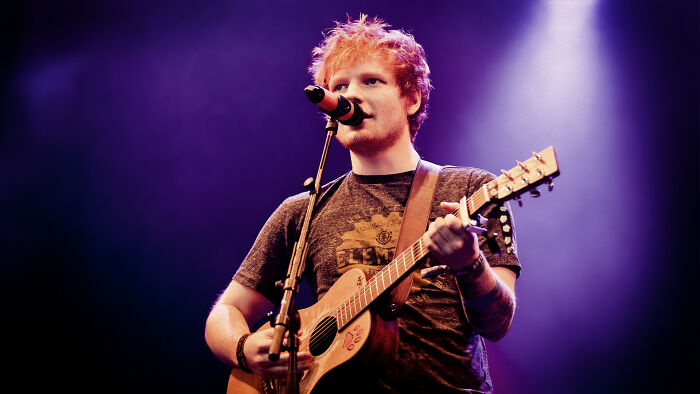 Ed Sheeran performing live with a guitar and microphone, bathed in purple light, embodying artistic comebacks.