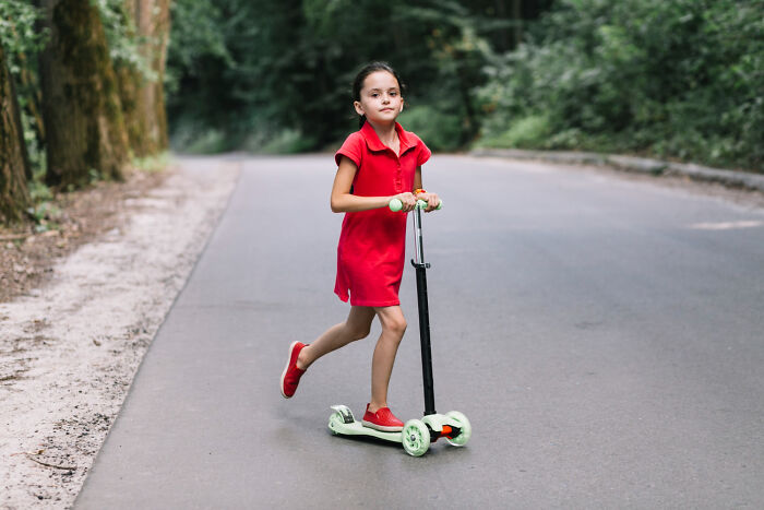 A young girl in a red dress rides a scooter on a paved path in a park, evoking a sense of childhood memories.
