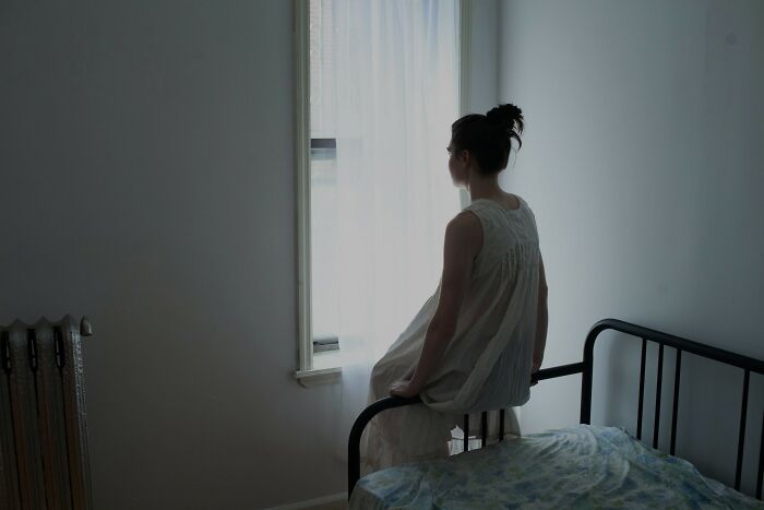 A person in a light-colored nightgown sits on a bed, gazing out a window, perhaps reflecting on unsettling childhood memories.