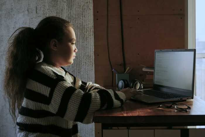 A young girl in a striped sweater sits at a desk, looking at a laptop, a scene that could trigger unsettling childhood memories.