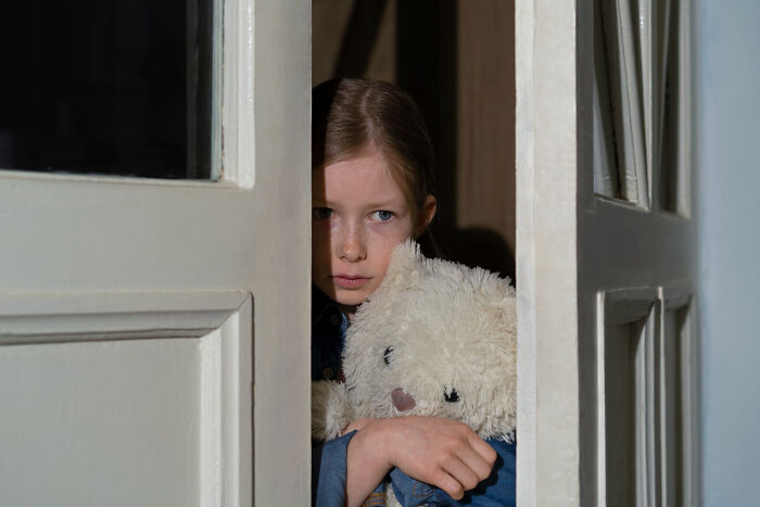 A young girl peeking through a doorway holding a teddy bear, reflecting unsettling childhood memories.