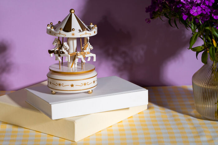 A gold and white carousel music box on white and yellow books, with a vase of purple flowers, representing childhood memories.